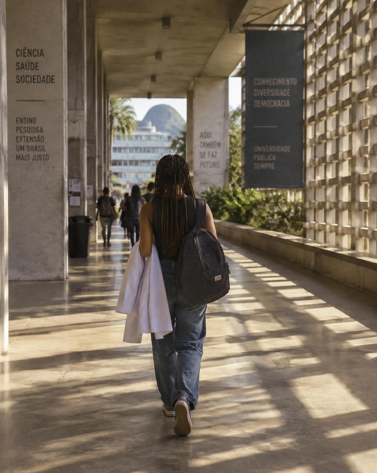 Estudante caminhando de costas por um corredor aberto de universidade, com sol da tarde no piso
