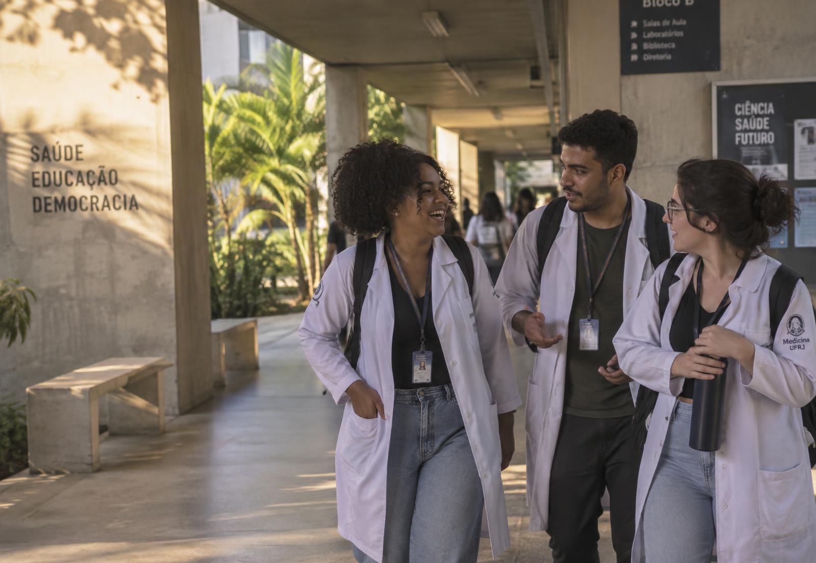 Três estudantes de Medicina conversando no corredor aberto de uma universidade brasileira, no fim da tarde